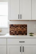 A rectangular wooden cutting board with a checkered block pattern in multiple shades of brown and tan wood, resting on a speckled granite countertop against a white wall.