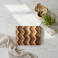 A square wooden cutting board with a chevron pattern of alternating light and dark wood stripes, resting on a speckled granite countertop.
