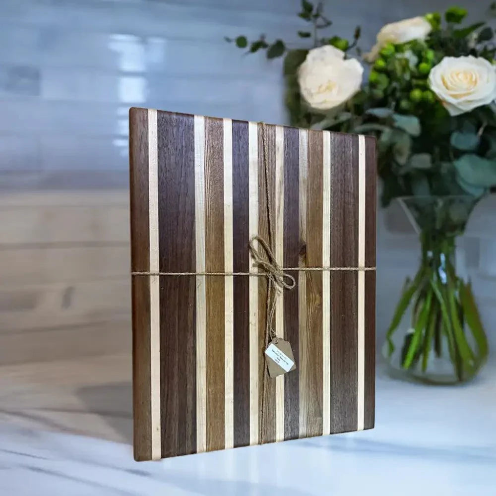 Handcrafted cutting board with stripes of Black Walnut, Maple, and Black Ash, 12"x15"x1.5", on marble countertop, with non-slip feet, in elegant kitchen setting.